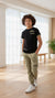 Child wearing a black t-shirt and green pants standing in a bright room with large windows.