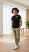 Child wearing a black t-shirt and green pants standing in a bright room with large windows.