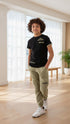 Child wearing a black t-shirt and green pants standing in a bright room with large windows.