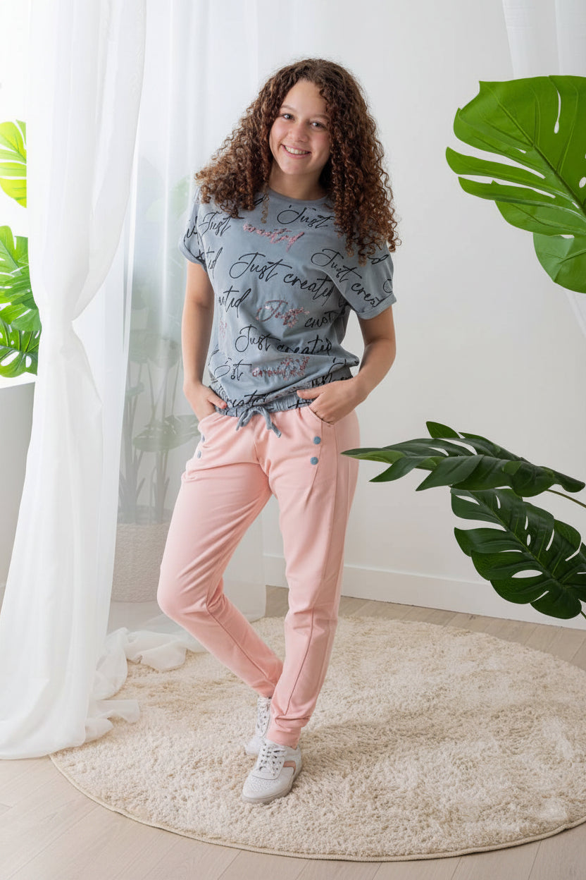 Girl wearing a gray t-shirt and pink pants standing in a room with plants and 'Rama Kids Wear' branding.