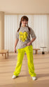 Young girl wearing a gray t-shirt with a yellow design and bright yellow pants, standing in a room with plants.